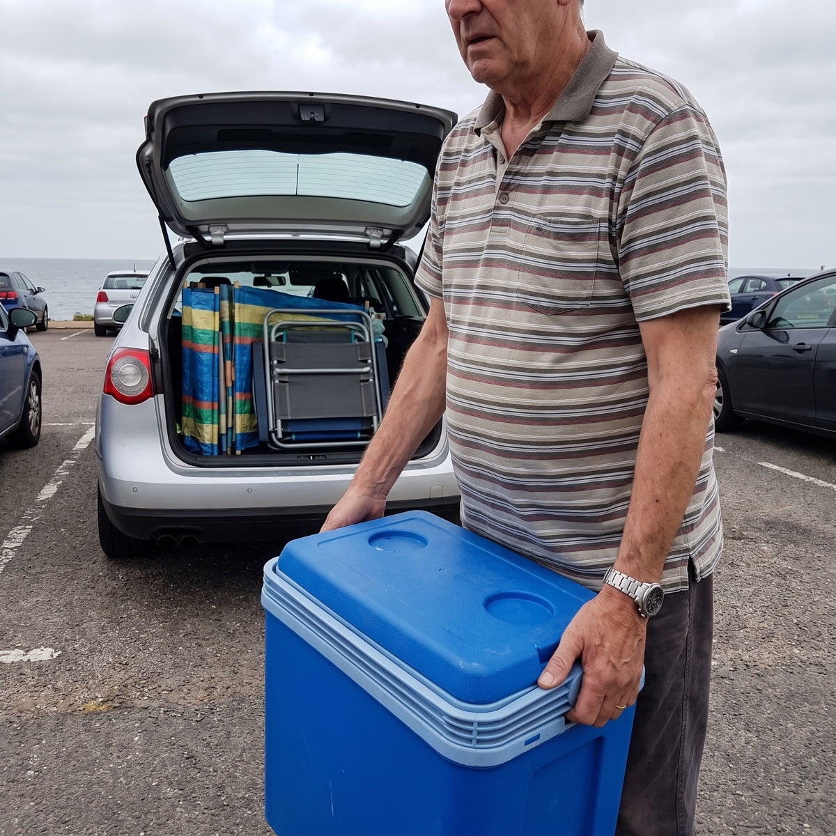 Man with thin arms and a round belly carrying a cool box